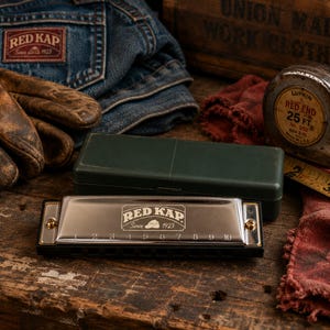 May include: A silver-colored harmonica with ten numbered holes and the text "RED KAP Since 1923" engraved on the front. The instrument is positioned in front of a dark green rectangular case, with a metal measuring tape showing 2 inches visible.