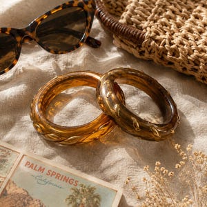 May include: Two translucent amber-colored resin bangles featuring a raised, braided gold-tone metal inlay. The bracelets are positioned near a pair of tortoiseshell cat-eye sunglasses, a woven straw bag, and a postcard with the text Palm Springs.