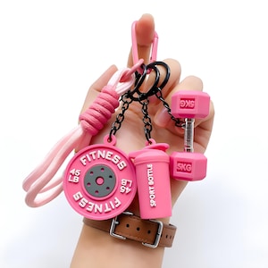Op de afbeelding: Een roze fitness-thema sleutelhanger met een halter van 5KG, een gewichtschijf van 45 LB en een sportfles. De sleutelhanger bevat ook een roze clip en een roze gevlochten polsband. De items zijn verbonden door zwarte kettingen.