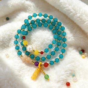 May include: A set of three turquoise beaded bracelets with gold accents. The bracelets feature a variety of colorful beads, including yellow, red, purple, and green, and a large white pearl. A yellow charm hangs from one bracelet.