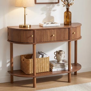 May include: A wooden console table with a half-moon design. It features three drawers with ribbed detailing and gold-colored knobs. The table has a lower shelf and is supported by rounded legs. A lamp, vase, and decorative items are displayed on the table.