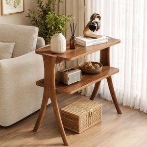 May include: A two-tiered wooden side table with a natural finish. The top shelf holds a white vase, a brown glass bottle, and books. The lower shelf features a small speaker and a wooden bowl. A woven basket sits below.
