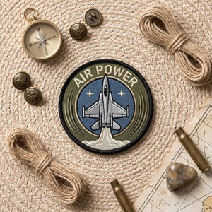 May include: A circular embroidered patch featuring a silver fighter jet graphic, the text AIR POWER, and a blue sky background with stars. The patch is surrounded by a brass compass, four metal star buttons, two coils of jute rope, and two brass casings.