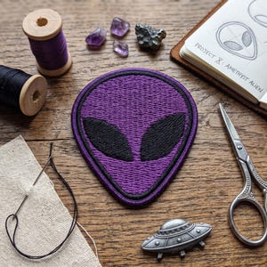 May include: A purple alien patch with black eyes and a black border sits on a wooden surface. The patch is surrounded by sewing supplies, including thread spools, a needle, and scissors. The text "PROJECT X: AMETHYST ALIEN" is visible in a notebook.
