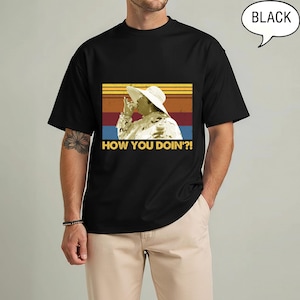 May include: Black t-shirt featuring a retro-style graphic with the phrase "HOW YOU DOIN'?!" and an image of a person wearing a white hat. The graphic has a vintage look with a sunset-like background. The word "BLACK" is in a speech bubble.
