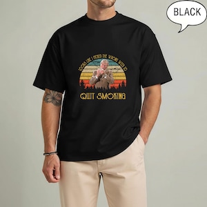 May include: Black t-shirt with a graphic that reads "Looks like I picked the wrong week to quit smoking." The graphic features a man in a suit, a sunset, and forest silhouette. The shirt is a classic crew neck.