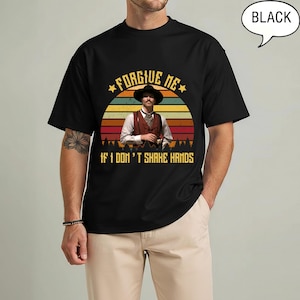 May include: Black t-shirt featuring a vintage-style graphic of a man in a cowboy hat and vest against a sunset backdrop. The text "FORGIVE ME" and "IF I DON'T SHAKE HANDS" are displayed above and below the image.