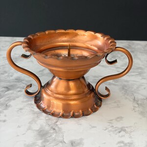 May include: A copper-colored metal candle holder featuring a wide, scalloped rim and a central spike for securing a candle. The base is flared with a matching scalloped edge, and two curved, scroll-shaped handles extend from the sides.