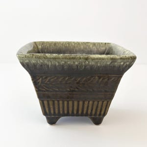 May include: A square ceramic planter with a dark brown base and a lighter, mottled green glaze along the top rim. The exterior features a textured band of leaf-like patterns above a series of vertical ridges, supported by four small, sturdy feet.