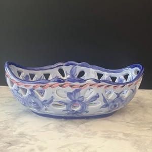 May include: An oval ceramic basket with a scalloped rim and openwork lattice sides. The white surface features hand-painted blue floral motifs and a decorative pink and black twisted rope pattern along the upper edge of the bowl structure.