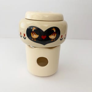 May include: A cream-colored ceramic oil warmer consisting of a base with a circular opening and a removable top bowl. The bowl features a black heart-shaped graphic containing two yellow ducks with red ribbons and a small red heart, flanked by red tulips.