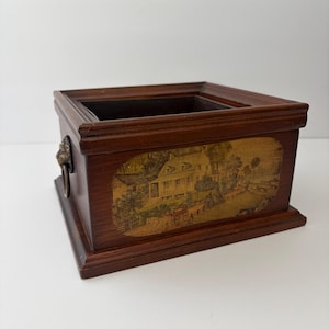 May include: A rectangular wooden box with a dark brown finish, featuring a decorative oval panel on the front that displays a printed illustration of a house and landscape. The box includes a metal lion head handle on the side and an open top.