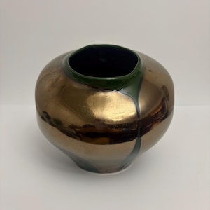 May include: A rounded ceramic vase with a metallic bronze finish and a smooth, dark green glazed rim. The glaze drips down the side of the vase in a single vertical line, contrasting with the reflective, warm-toned surface of the main body.