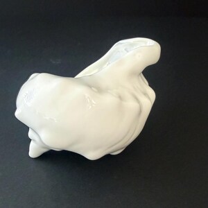 May include: A white ceramic vessel with an irregular, organic, and sculptural shape. The surface is smooth and glossy, featuring deep indentations and rounded protrusions that create a fluid, asymmetrical form with a single open top aperture.