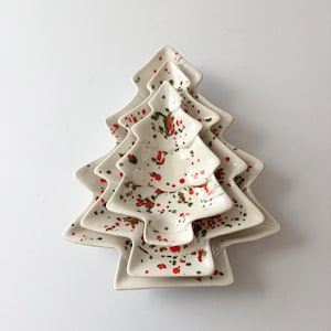 May include: A set of three nested ceramic dishes shaped like Christmas trees. Each white dish features a speckled pattern of small red and green paint splatters across the surface, creating a festive look for these serving pieces.