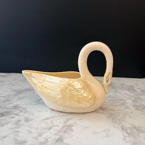 May include: A cream-colored ceramic swan-shaped dish with a glossy, iridescent finish. The swan features a curved neck, a defined beak with a small black eye detail, and molded wing textures on the sides of the body, creating a hollow vessel.