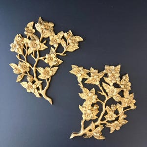 May include: Two gold-colored wall plaques featuring a relief design of flowering branches with detailed blossoms and leaves. The metallic finish has a reflective quality, highlighting the intricate textures of the petals and stems on each piece.