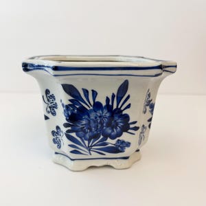 May include: A hexagonal ceramic planter featuring a white base with blue floral and butterfly patterns. The rim and base edges are accented with blue lines, and the design includes detailed blue blossoms and small flying butterflies.