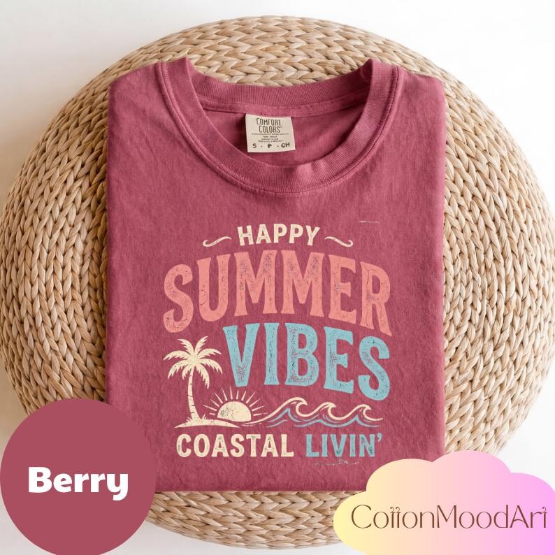 May include: Berry-colored t-shirt with the words "HAPPY SUMMER VIBES COASTAL LIVIN'" in a retro font. The design includes a palm tree, sun, and waves. The shirt is folded on a woven surface.