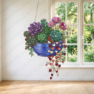May include: A stained glass suncatcher featuring a blue bowl filled with green and purple succulents, pink flowers, and a trailing string of heart-shaped leaves in shades of red and orange, suspended by a silver metal chain against a window.
