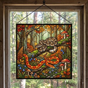 May include: A square stained glass panel hanging by a black chain in a window. The artwork depicts a forest scene with a brown patterned snake coiled on a branch above a bright orange and yellow snake, surrounded by mushrooms and blue flowers.