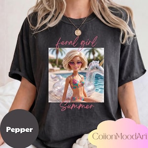 May include: Pepper-colored t-shirt featuring a graphic of a Barbie doll in a bikini, floating on a unicorn pool float. The words "feral girl" and "Summer" are written in pink script. The shirt has a relaxed fit.
