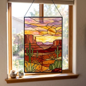 May include: A rectangular stained glass panel featuring a desert landscape with saguaro cacti, barrel cacti, and agave plants under a sunset sky. The scene includes mesas and rock formations in shades of orange, red, yellow, and purple, framed in black.