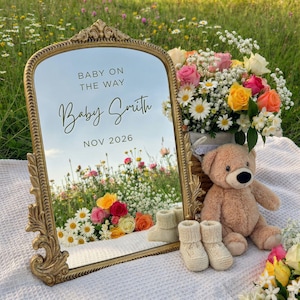 Pode incluir: Um espelho dourado com o texto "BABY ON THE WAY Baby Smith NOV 2026" é rodeado por flores, um ursinho de pelúcia e botinhas de bebê. A cena é ao ar livre com um campo de flores ao fundo, anunciando um nascimento.