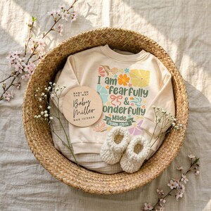 Pode incluir: Um suéter bege para bebê com o texto "I am fearfully & wonderfully made" e gráficos florais, dentro de uma cesta de vime. Uma placa de madeira diz "Baby Miller Due 12/202X". Botinhas de bebê brancas de crochê também estão na cesta.