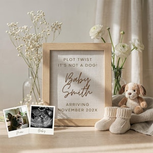 Puede incluir: Anuncio enmarcado con el texto "PLOT TWIST IT'S NOT A DOG! Baby Smith ARRIVING NOVEMBER 202X." El marco está junto a una foto de un perro y una ecografía. Un cachorro de peluche, patucos y flores completan la escena.