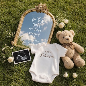 Op de afbeelding: Een aankondigingsscène op groen gras met een goudomrande spiegel met tekst, een witte romper met "Baby Robinson", een teddybeer en een echo. De spiegel weerspiegelt een blauwe lucht met wolken en de woorden "for this child we have prayed".