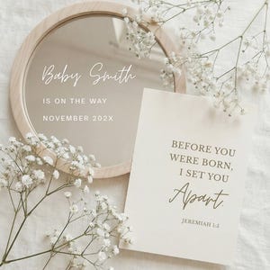 Pode incluir: Um espelho redondo com moldura de madeira clara, com o texto "Baby Smith IS ON THE WAY NOVEMBER 202X". Um cartão com o texto "BEFORE YOU WERE BORN, I SET YOU Apart JEREMIAH 1:5" está ao lado do espelho. Flores brancas dispostas ao redor do espelho e do cartão.