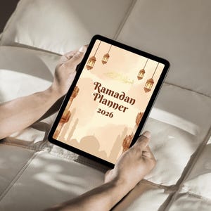 May include: A digital tablet displaying a "Ramadan Planner 2026" design. The planner features a warm color palette with illustrations of lanterns and a mosque silhouette. The tablet is held by hands, suggesting a user-friendly interface.