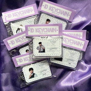 May include: A collection of ID keychains, each in a clear plastic wrapper, rests on a purple satin background. The keychains display a photo and text. Each package has a lavender label with the text "ID KEYCHAIN ARBANG EDITION".