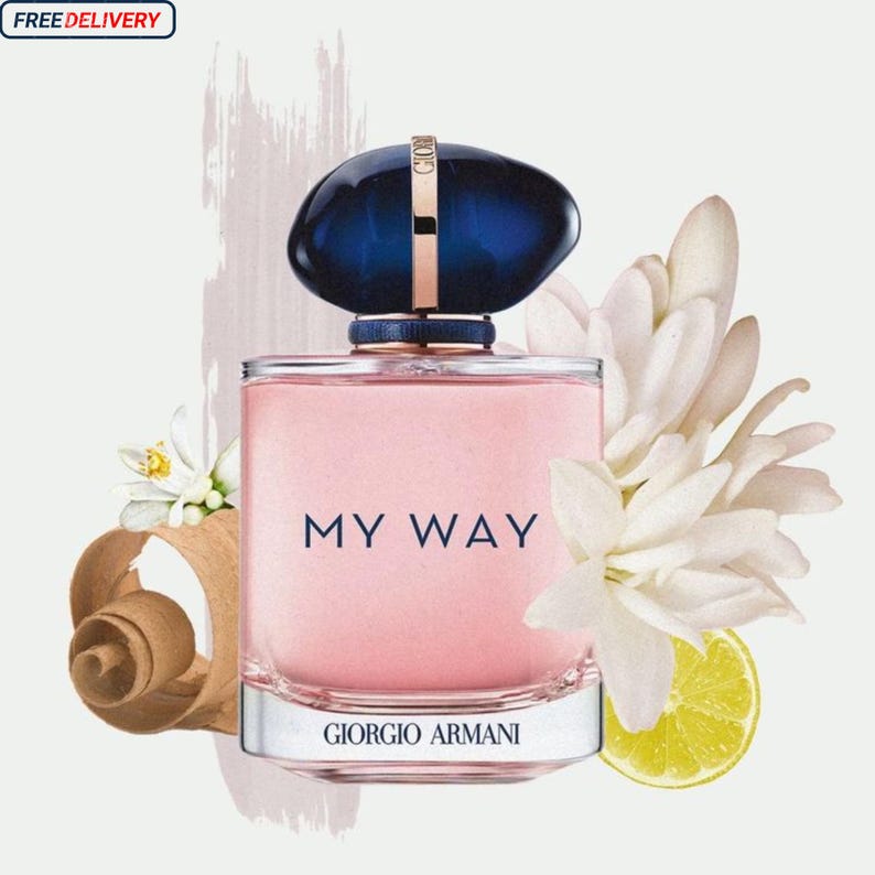 May include: A pink rectangular perfume bottle with a dark blue cap and the words "MY WAY" printed on the front. The bottle is surrounded by white flowers, a lemon slice, and wood shavings. The brand name "GIORGIO ARMANI" is printed at the bottom.