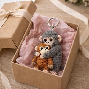 May include: A handmade crochet monkey keychain in a gift box. The gray monkey hugs a smaller brown monkey. The gift box is brown with a cream-colored ribbon and pink tissue paper. A charming and unique gift idea.