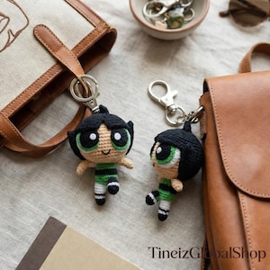 May include: Two crochet keychains featuring a cartoon character with black hair, green eyes, and a green and black outfit. The keychains are attached to silver metal clasps. The background includes a tan tote bag, a brown leather purse, and a small bowl.