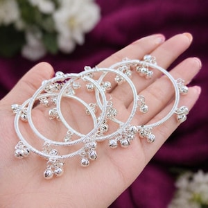 May include: Four silver anklets with dangling bells are displayed. The anklets are circular and have a textured design. The bells are small and round, adding a delicate touch. The anklets are shown on a hand against a dark purple background.