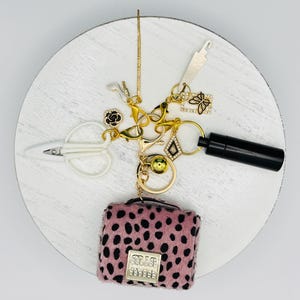 May include: A gold-toned keychain featuring a pink and black leopard print miniature handbag charm, a white scissor charm, a black cylindrical container, a butterfly charm, a high-heeled shoe charm, a black rose charm, and a gold sphere charm.