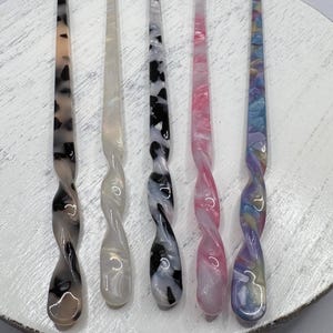 Könnte beinhalten: Fünf lange Haarstäbe aus poliertem Acryl mit einem glatten, gedrehten Design am unteren Ende. Die Stäbe weisen verschiedene Muster auf, darunter Schildpatt, cremefarben, schwarz-weiß marmoriert, rosa Wirbel und eine bunte Pastellmischung.