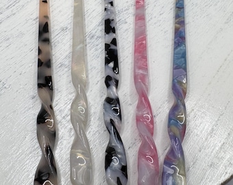 Könnte beinhalten: Fünf lange Haarstäbe aus poliertem Acryl mit einem glatten, gedrehten Design am unteren Ende. Die Stäbe weisen verschiedene Muster auf, darunter Schildpatt, cremefarben, schwarz-weiß marmoriert, rosa Wirbel und eine bunte Pastellmischung.