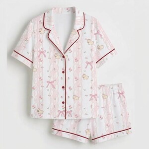 May include: A white pajama set with a pink and red pattern. The short-sleeved top and shorts feature a repeating design of pink bows, flowers, and small animal illustrations. The edges are trimmed with a dark red piping.