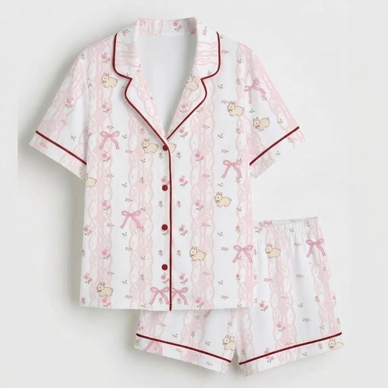 May include: A white pajama set with a pink and red pattern. The short-sleeved top and shorts feature a repeating design of pink bows, flowers, and small animal illustrations. The edges are trimmed with a dark red piping.