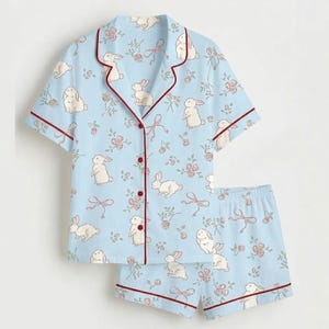 May include: Pale blue pyjama set featuring a pattern of white rabbits, pink flowers, and bows. The short-sleeved top and shorts are trimmed with burgundy piping. The button-down top has a notched collar.