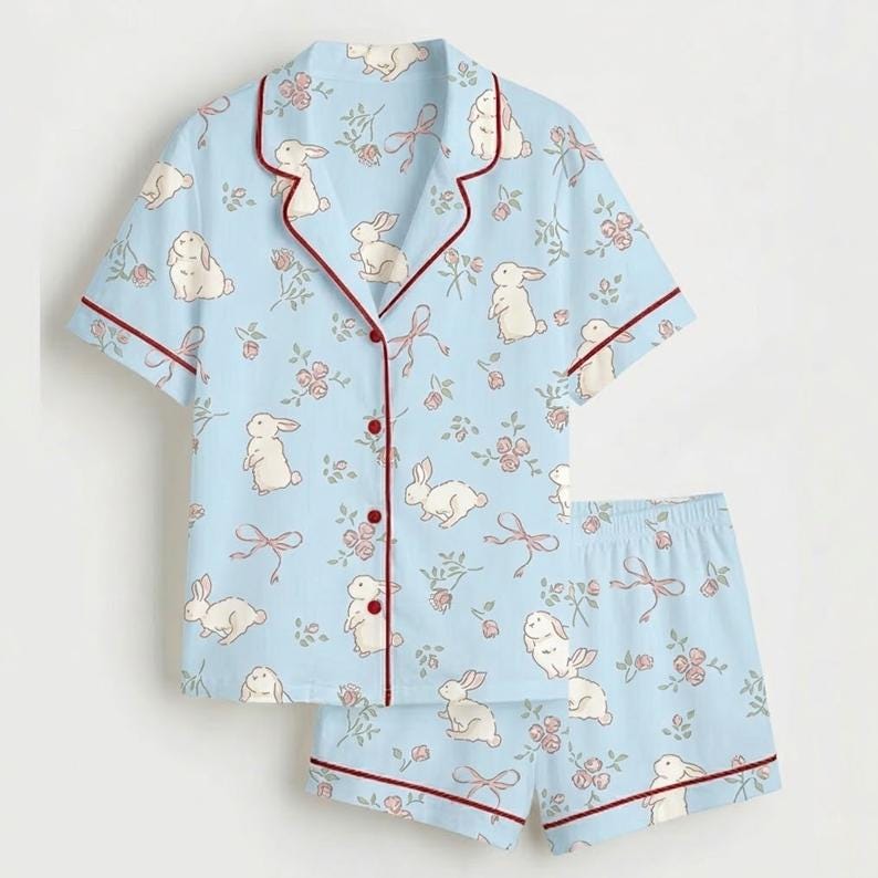May include: Pale blue pyjama set featuring a pattern of white rabbits, pink flowers, and bows. The short-sleeved top and shorts are trimmed with burgundy piping. The button-down top has a notched collar.