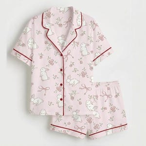 May include: A light pink pajama set with a pattern of white rabbits, pink roses, and bows. The short-sleeved top and shorts have burgundy trim. The set is made of a soft, lightweight fabric.
