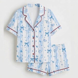 May include: A two-piece pyjama set with a short-sleeved button-down shirt and matching shorts. The set is white with a blue floral and bow pattern, accented with burgundy trim.
