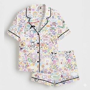 May include: White pajama set with a colorful, whimsical print of cartoon characters, food, and text. The short-sleeved top and shorts are outlined in black. The word "HAPPY" is visible on the top.