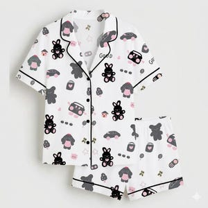 May include: White pajama set with a playful pattern of cartoon bunnies, bears, cars, and other small icons. The short-sleeved top and shorts are trimmed with black piping. The word "Good" is printed on the top.