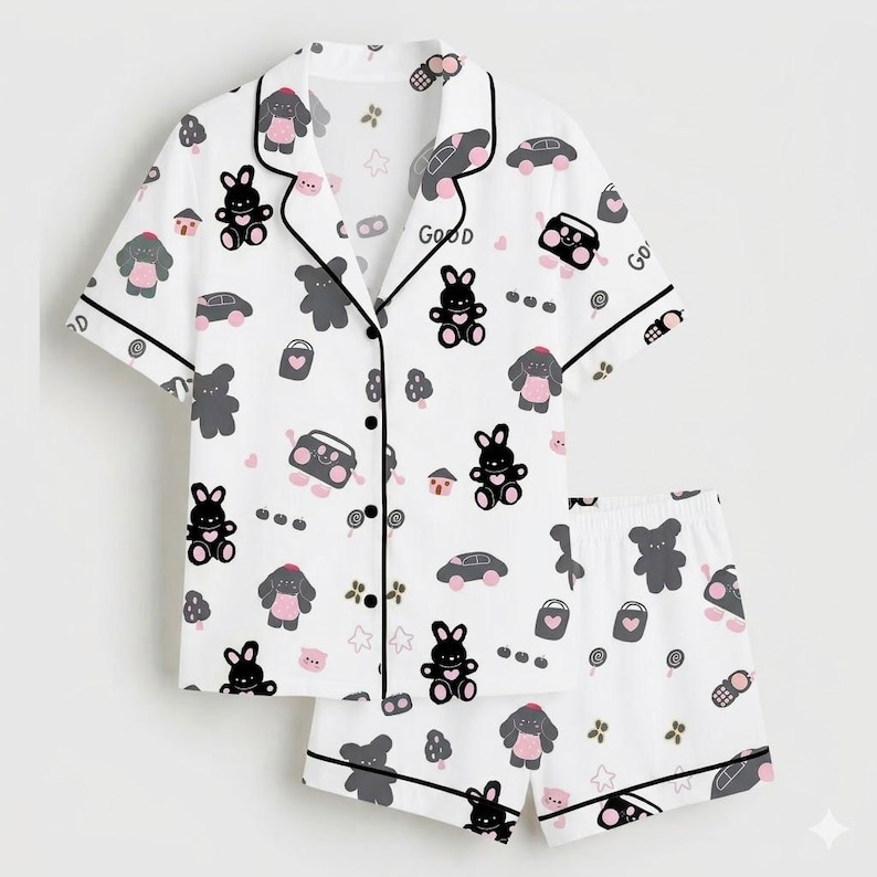 May include: White pajama set with a playful pattern of cartoon bunnies, bears, cars, and other small icons. The short-sleeved top and shorts are trimmed with black piping. The word "Good" is printed on the top.